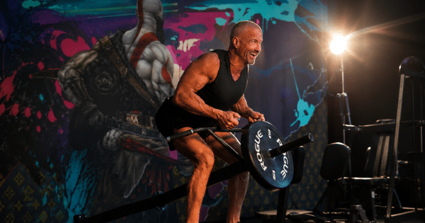 A muscular, fit man in a black tank top smiles while performing a bent-over row exercise with a T-bar row machine in a gym setting.