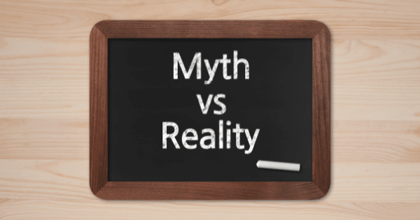 A wooden-framed miniature chalkboard resting on a light wood surface displays the words "Myth VS Reality" written in white chalk, with a piece of chalk lying beside it.