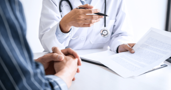 A doctor in a white lab coat points a pen toward a document held during a consultation with a patient seated across the desk.