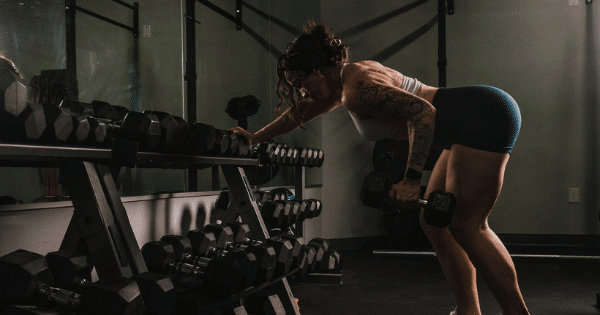 A tattooed woman in a sports crop top and shorts bends forward next to a dumbbell rack in a gym, performing a single-arm dumbbell row exercise.