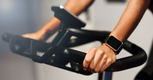 A close-up view of a person’s hands gripping the handlebars of a stationary exercise bike, wearing a rectangular black smartwatch on their left wrist.