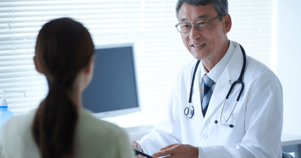 An older male doctor wearing glasses, a white lab coat, and a stethoscope speaks warmly to a patient seated across from him.