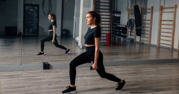 A woman in black athletic wear performs a walking lunge while holding light dumbbells in a mirrored gym studio.