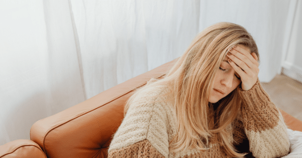 A blonde woman wearing a cozy sweater sitting on a brown leather couch with her hand on her forehead, looking fatigued or unwell.