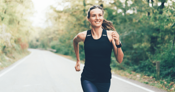 A smiling woman with her hair tied back jogging down a paved road framed by lush green trees, dressed in a black athletic tank top and dark leggings.