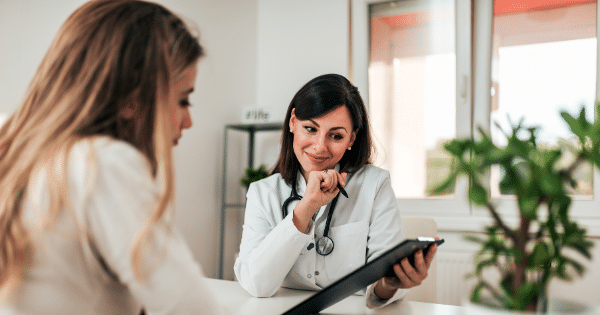 A female doctor holding a clipboard with a pen in hand, listening attentively to a patient during a consultation.