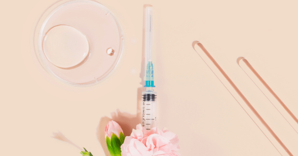 A flat lay arrangement showing a medical syringe with pink flowers, test tubes, and a Petri dish on a neutral background.