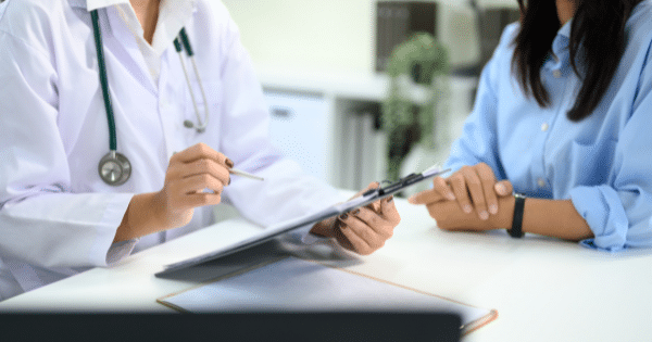 A doctor in a white coat explaining details from a chart or document on a clipboard to a patient seated across the desk.