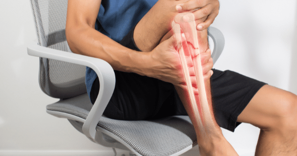 A person sitting in a mesh office chair clutching their lower leg, featuring a semi-transparent anatomical overlay highlighting a shin bone fracture with red pain indicators.