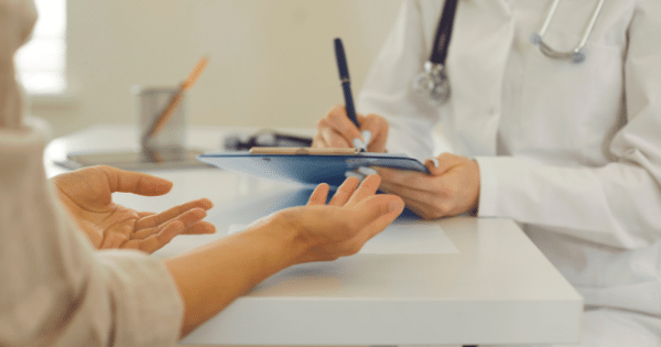A close-up of a doctor writing notes on a clipboard while a patient across the desk gestures with open hands during a consultation.