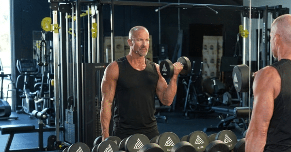 A fit, muscular middle-aged man in a black tank top performing bicep curls with a dumbbell in front of a gym mirror.