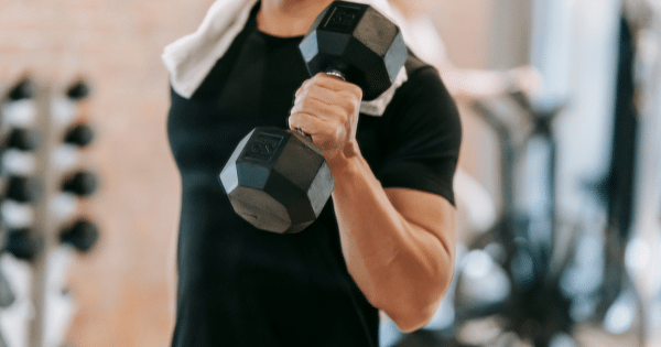 A close-up view of a person with a white towel draped over their shoulders performing a single-arm dumbbell bicep curl in a gym.