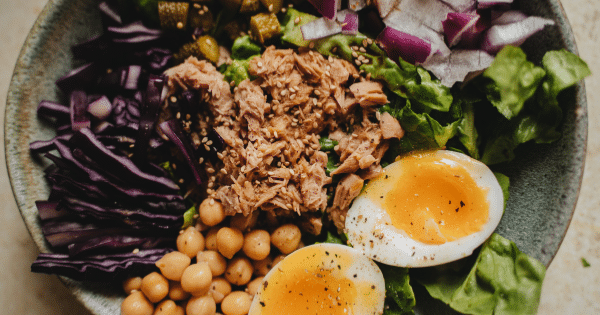An overhead view of a healthy salad bowl filled with shredded tuna, two soft-boiled egg halves, chickpeas, shredded red cabbage, fresh green lettuce, chopped red onions, and sesame seeds.