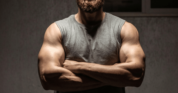 A muscular man with a full brown beard standing in dramatic overhead lighting with his arms crossed over a grey sleeveless top.