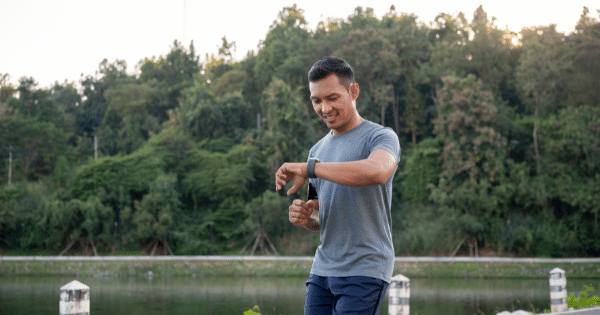 A man in a grey t-shirt standing outdoors near a body of water and dense forest, looking down at his smartwatch with a smile.