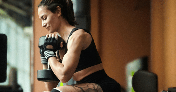 A fit woman wearing workout gloves and a black crop top performing a seated concentration curl with a heavy dumbbell in a gym.
