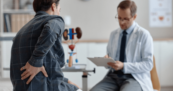 A male patient holding his lower back while speaking with a doctor in a white lab coat, with an anatomical model of the urinary system sitting on the desk between them.