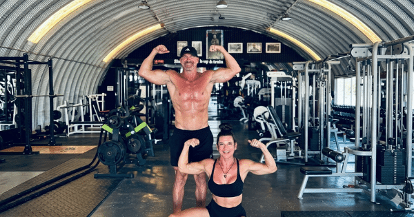 fit man wearing a black cap and shorts flexing his biceps alongside a smiling woman in a black sports bra kneeling in front of him inside a Quonset hut gym facility.