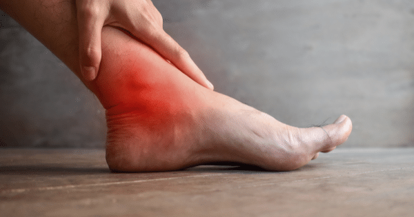 Side view of a human foot and ankle on a wooden floor, with a highlighted red glowing area on the ankle joint indicating pain or inflammation.