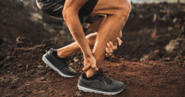A runner crouched on a rocky trail holding their injured lower leg and ankle.