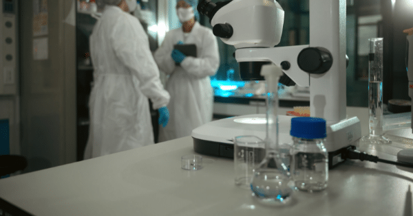 Laboratory setting with glassware and a white microscope on a table, with blurred scientists in protective suits in the background.