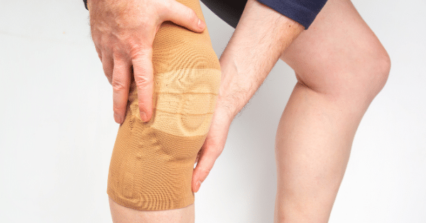Close-up of a person wearing a tan elastic support sleeve on their knee while holding it with both hands.