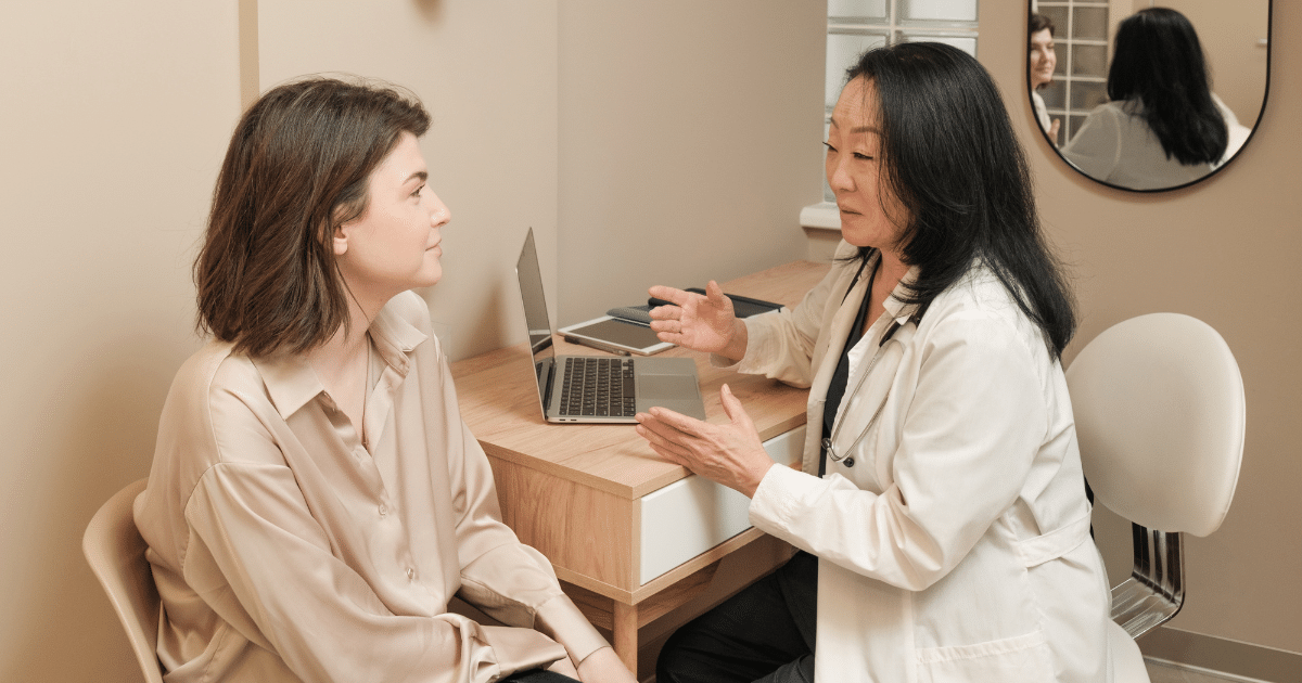 A female doctor in a white lab coat sitting at a wooden desk with a laptop, gesturing with her hands while speaking to a patient sitting opposite her.