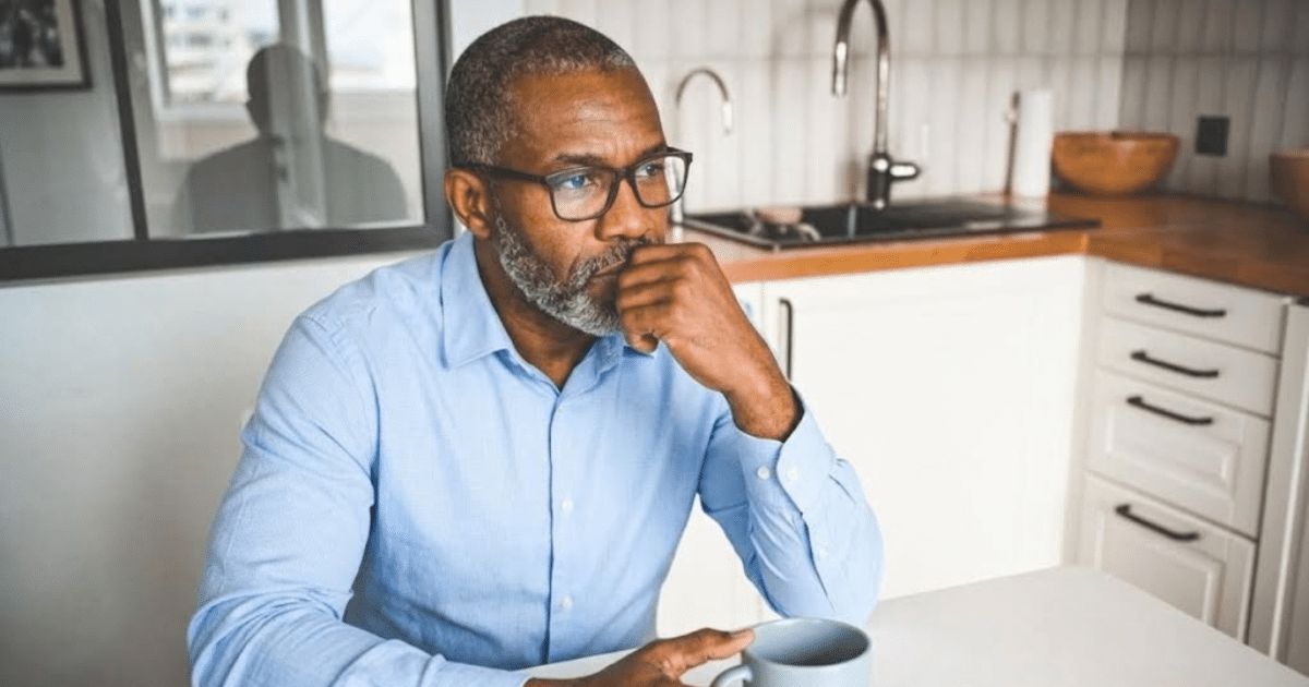 A middle-aged Black man wearing glasses and a light blue dress shirt sitting thoughtfully at a table in a bright kitchen, resting his chin on his hand next to a coffee mug.