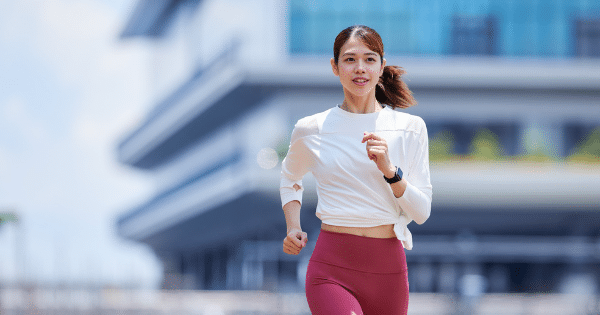 young woman with a ponytail jogging outdoors in a white long-sleeve athletic top, red leggings, and a smartwatch, framed against a modern blurred building.