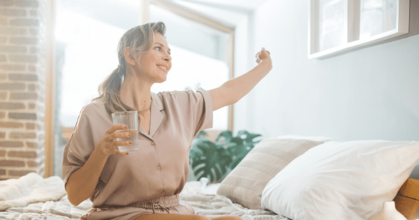 A smiling woman in beige silk pajamas sitting on a bed, holding a glass of water in one hand and stretching her other arm outward in a brightly lit room.