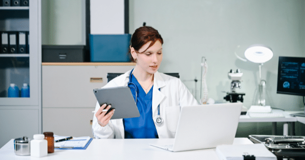 A female doctor wearing blue scrubs and a white lab coat sitting at a desk with a stethoscope around her neck, looking at a digital tablet while working next to a laptop.