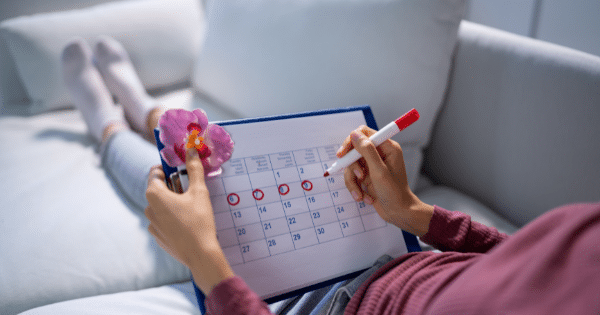 An overhead shot of a person relaxing on a white couch with legs elevated, using a red marker to circle dates on a monthly calendar held on a clipboard.