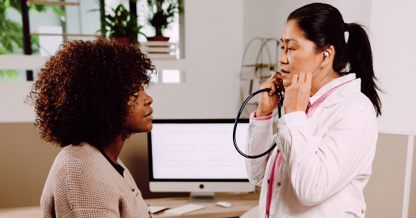 A female doctor wearing a stethoscope around her neck speaking with a patient in a consultation room with a computer monitor in the background.