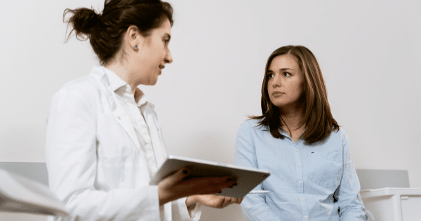 A female doctor in a white coat holding a digital tablet and talking to a patient dressed in a light blue button-down shirt.