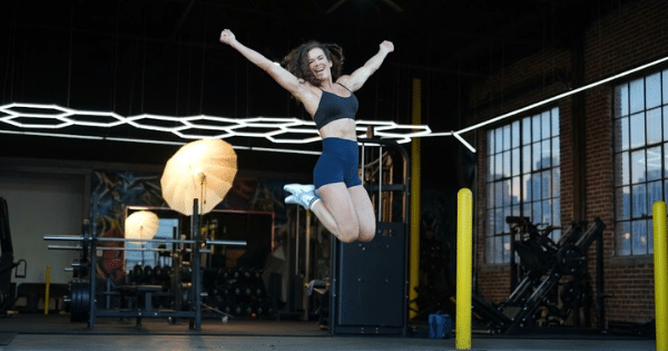 An athletic woman in a black sports bra and blue shorts jumping high into the air with arms outstretched and a joyful expression inside a spacious gym setup.