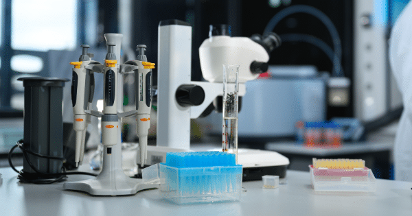 Laboratory counter equipped with lab pipettes, tip boxes, a graduated cylinder, and a microscope.