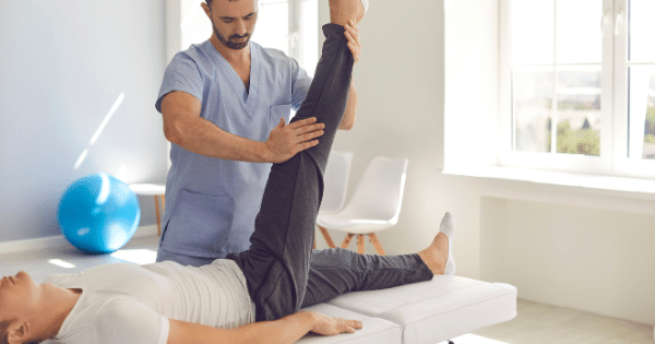 A male physical therapist in blue scrubs lifting and stretching a male patient's leg on an examination table.