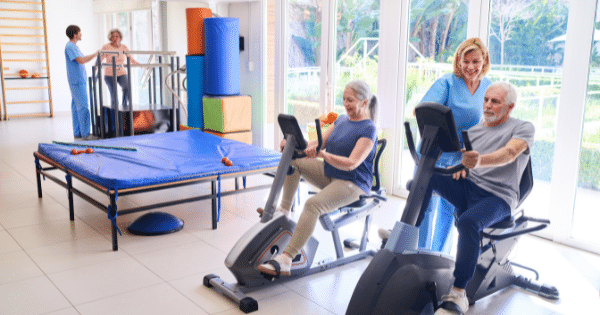 Senior individuals exercising on recumbent stationary bikes in a brightly lit rehabilitation center with assistance from physical therapists.