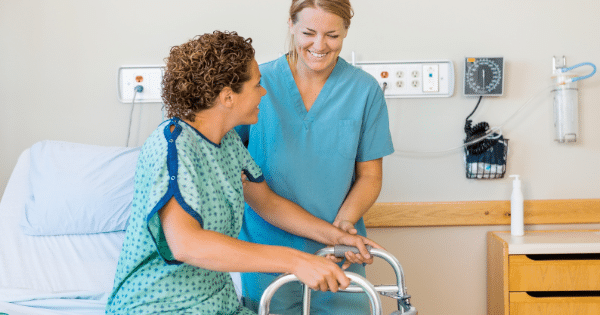 A female nurse in blue scrubs smiling while helping a patient in a hospital gown stand up using a walker next to a hospital bed.