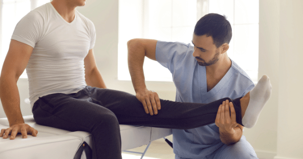 A physical therapist guiding a male patient through a leg extension exercise while sitting on an examination table.