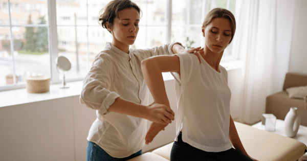A female therapist performing a posture and shoulder adjustment on a female patient sitting on a therapy bench in a sunlit room.