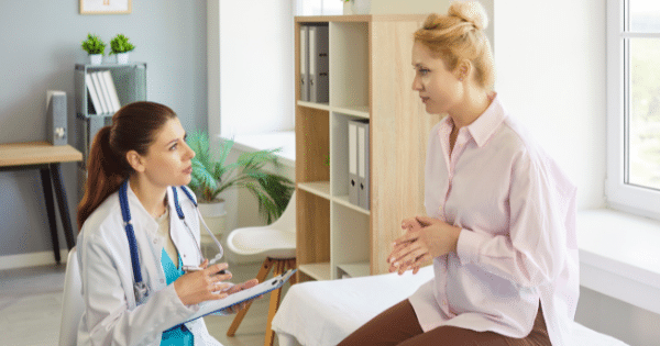 A female doctor in a white lab coat taking notes on a clipboard while speaking with a female patient seated across from her in a brightly lit medical office.