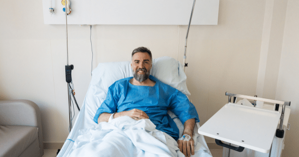 A smiling middle-aged man with a beard wearing a blue hospital gown resting in an adjustable hospital bed with an IV line attached to his hand.