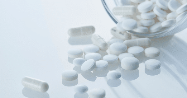 A close-up view of various white pharmaceutical pills and capsules spilling out of a small glass bowl onto a reflective white surface.