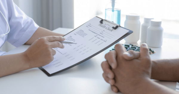 A doctor holding a pen pointing to a "Health Check" form on a clipboard while seated across a desk from a patient with clasped hands.