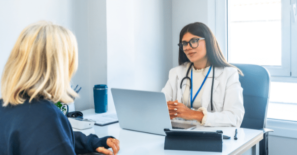 A female doctor wearing glasses, a white coat, and a stethoscope talking to a blonde female patient across a desk with a laptop open between them.