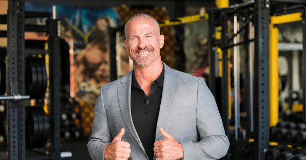 smiling man in a grey suit jacket giving a double thumbs-up inside a gym setting.