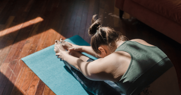 A high-angle view of a woman sitting on a yoga mat and stretching forward to reach her feet in a sunlit room.