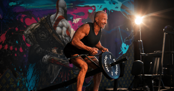 A smiling muscular man performing T-bar rows on a gym machine, positioned in front of a colorful wall mural.