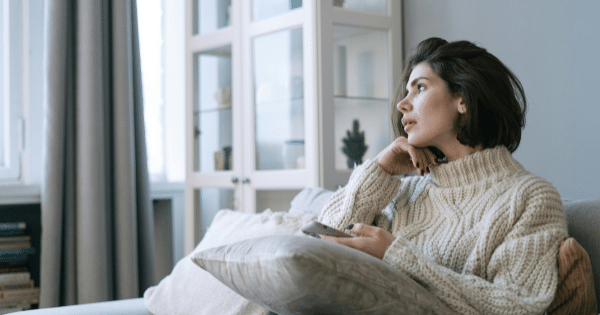 A woman wearing a cozy knit sweater sitting thoughtfully on a couch while holding her smartphone.